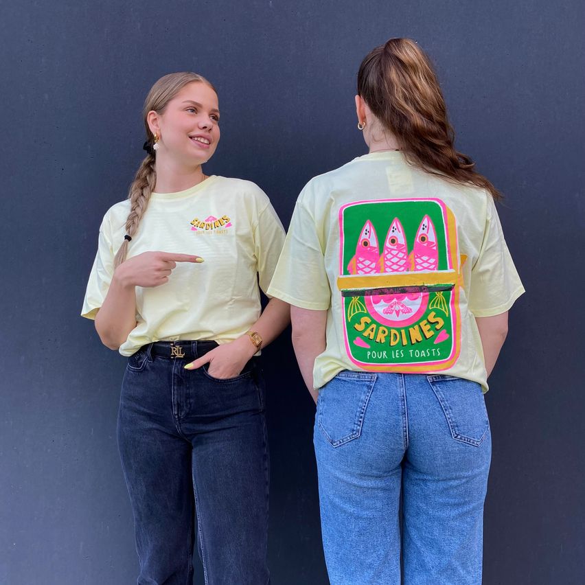 Zwei Frauen stehen nebeneinander und lächeln für ein Foto. Eine trägt ein gelbes T-Shirt mit einem Fisch-Grafik, die andere ein gelbes T-Shirt mit einem farbenfrohen Fisch-Grafik auf dem Rücken. Sie tragen beide Jeans und stehen vor einer grauen Wand.