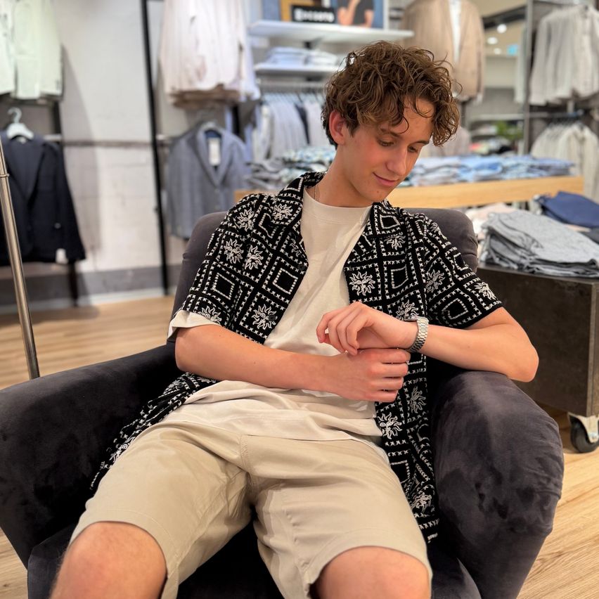 A young man with curly hair is sitting on a chair in a clothing store, looking at his watch. He is wearing a white shirt, beige shorts, and a patterned jacket.