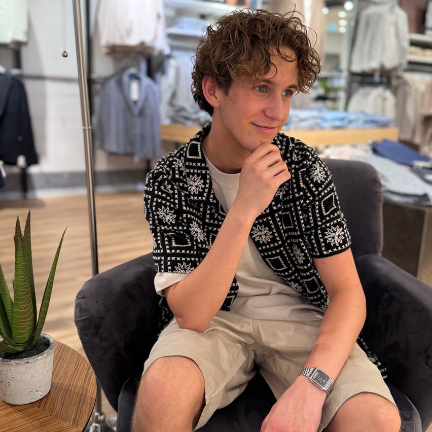 A young man with curly hair is sitting on a chair in a clothing store, smiling and posing for a photo. He wears a patterned shirt and khaki shorts. A potted plant is on the table beside him.