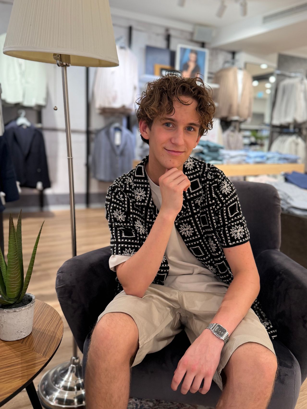A young man with curly hair is sitting in a chair in a clothing store, wearing a patterned shirt and khaki shorts, with a potted plant and a lamp in the background.
