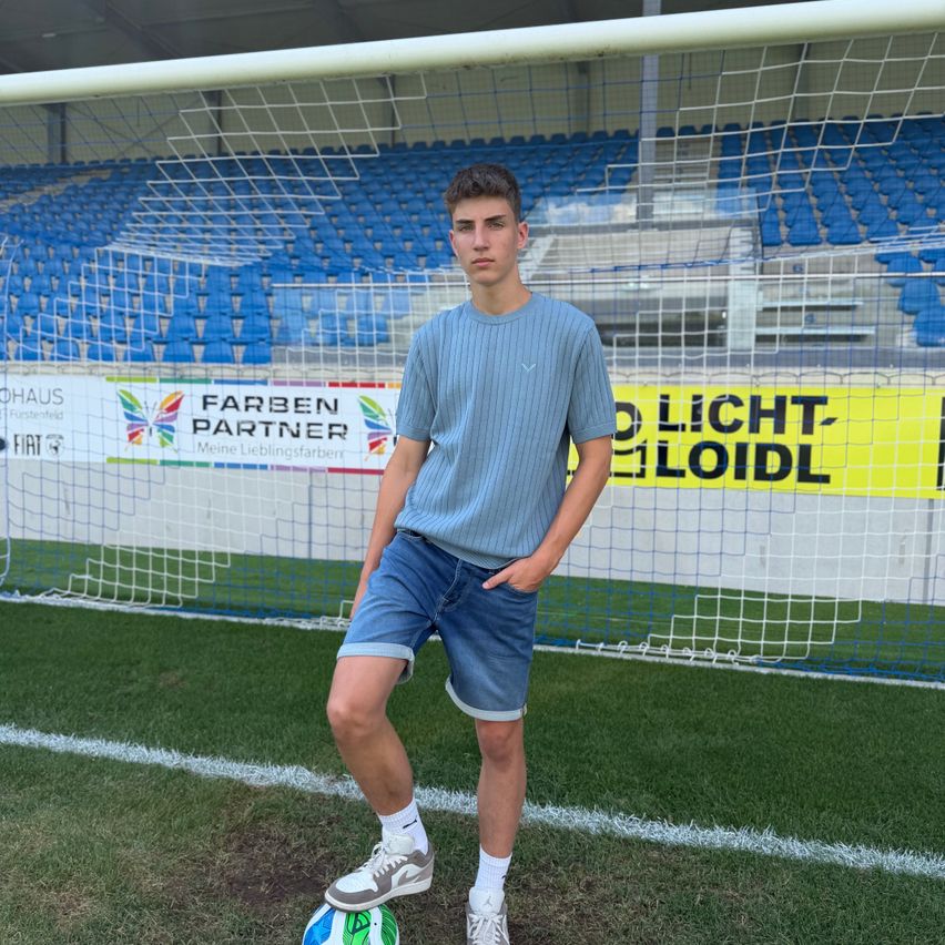Ein junger Mann steht auf einem Fußballfeld vor einem Tor, trägt ein hellblaues T-Shirt, Jeansshorts und weiße Socken. Er hat einen Fußball an seinen Füßen.