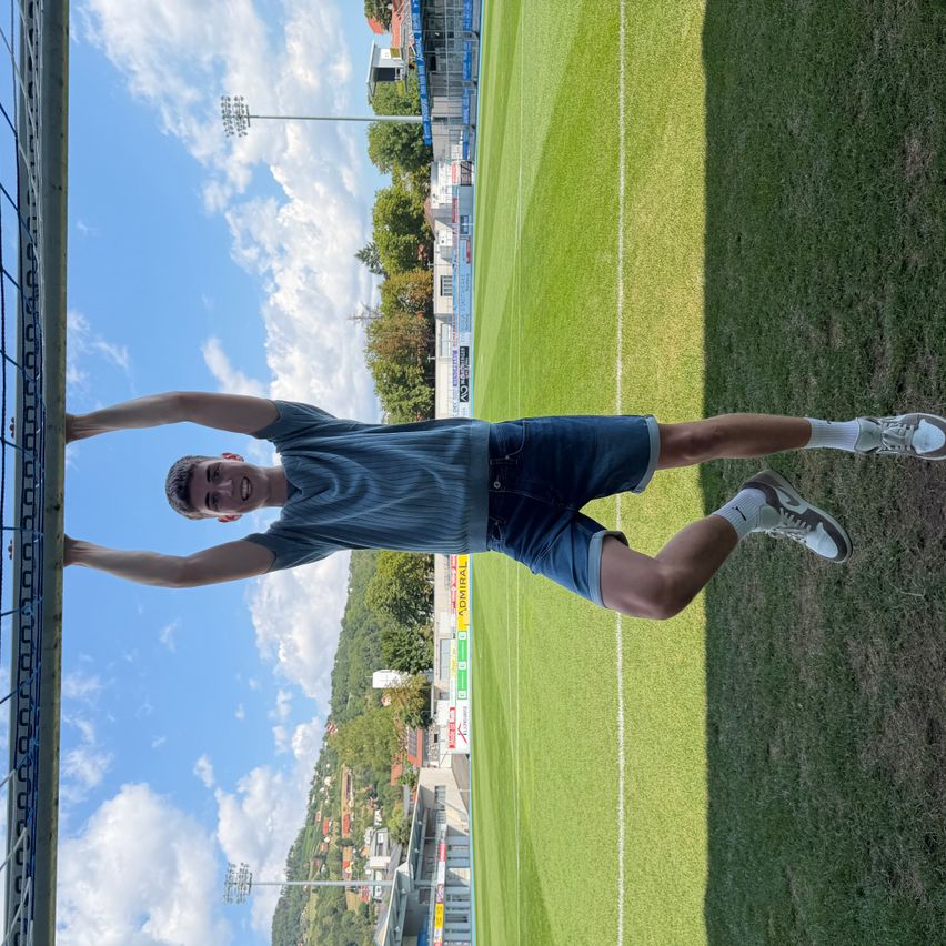 Ein junger Mann steht auf einem Fußballfeld, lehnt sich gegen einen Zaun und lächelt für ein Foto.