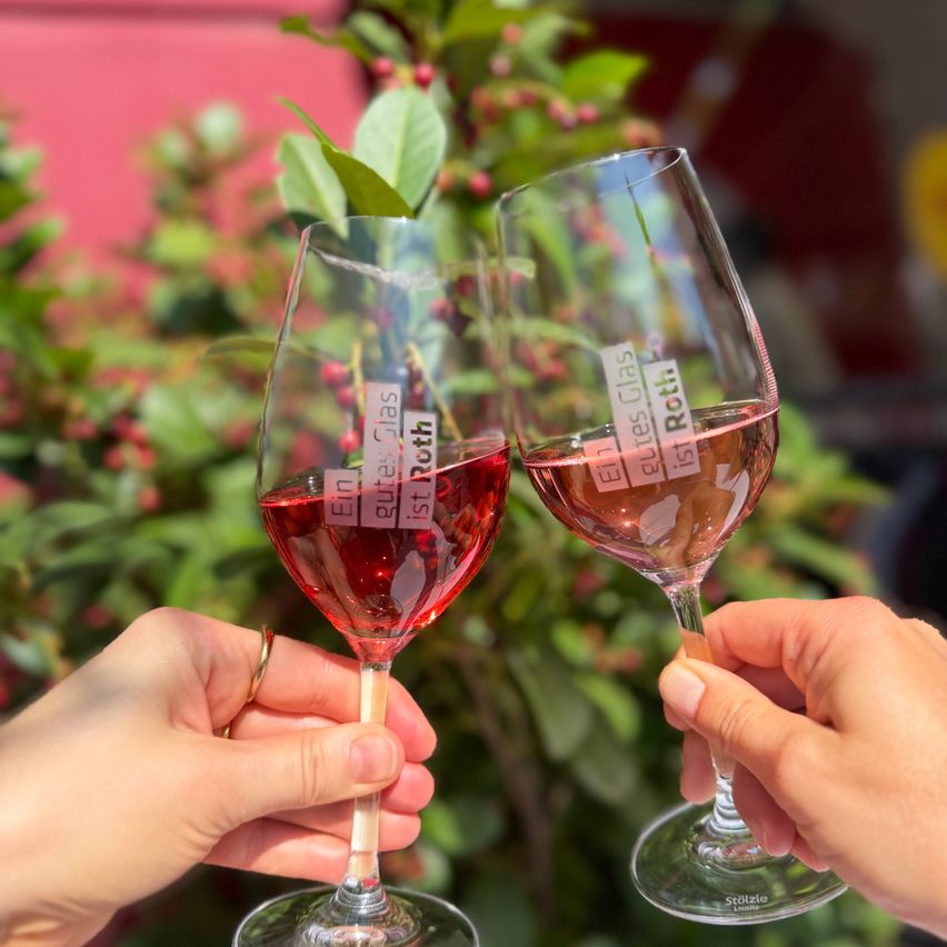 Zwei Hände halten Weingläser mit rotem Wein, eines mit der Aufschrift 'Ein Glas ist Ruth' und das andere 'Ein Glas ist Roth', vor einem unscharfen Hintergrund aus grünen Blättern.