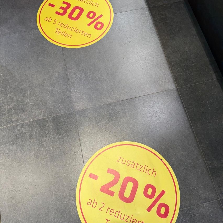 Zwei gelbe runde Schilder auf einem gefliesten Boden, eines zeigt '-30%' und das andere 'zusätzlich -20%'. Beide Schilder haben einen roten Rand und weißen Text.