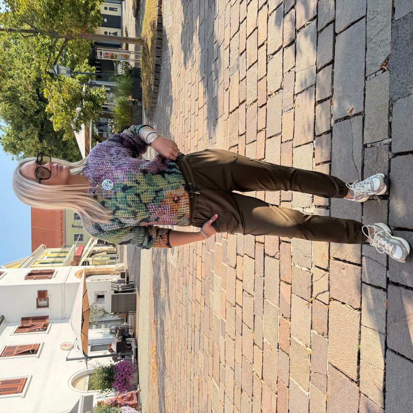 A woman with blonde hair is standing on a brick sidewalk, wearing a colorful sweater, brown pants, and white sneakers. She is posing for a photo with a building and trees in the background.
