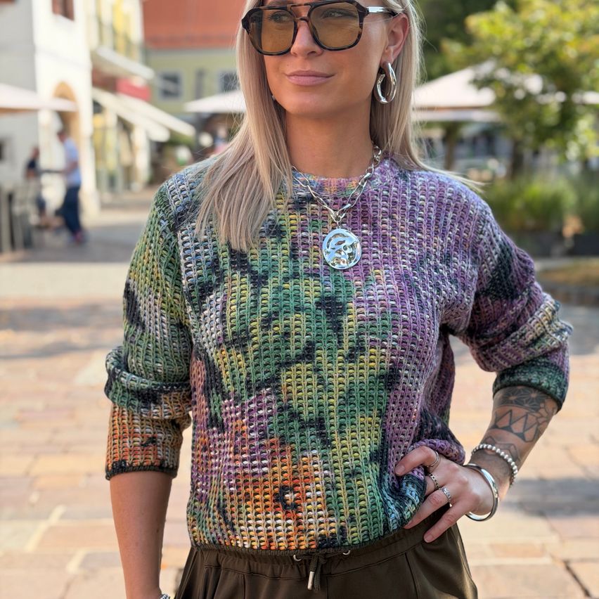 A woman with blonde hair wearing a colorful knitted sweater with a large pendant necklace stands on a cobblestone street.