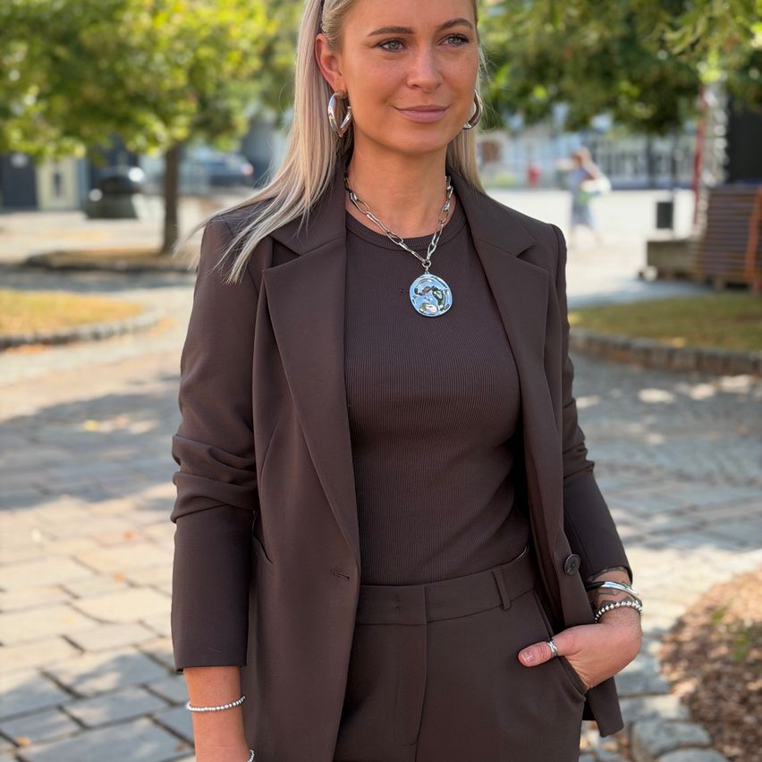 Eine Frau mit blonden Haaren, die eine braune Jacke und eine Halskette trägt, steht auf einem gepflasterten Weg. Hinter ihr sind Bäume, ein Gebäude und eine Person, die geht.
