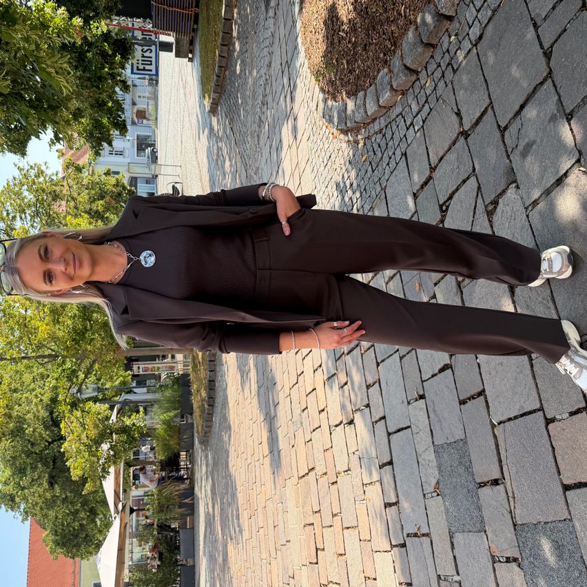 Eine Frau in einem braunen Anzug steht auf einem gepflasterten Gehweg und posiert für ein Foto. Hinter ihr befinden sich Bäume und ein Gebäude mit einem Schild, das 'Fürst' steht.