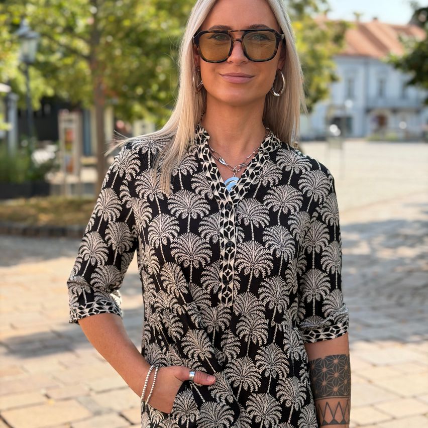 A woman with blonde hair wearing a black and white palm tree print shirt stands on a cobblestone street.