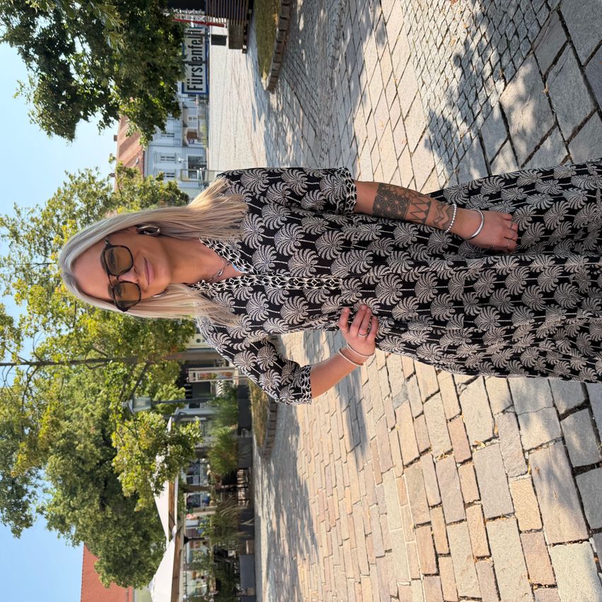 A woman with blonde hair and a tattoo on her arm is standing on a brick sidewalk, wearing a black and white patterned dress and sunglasses.