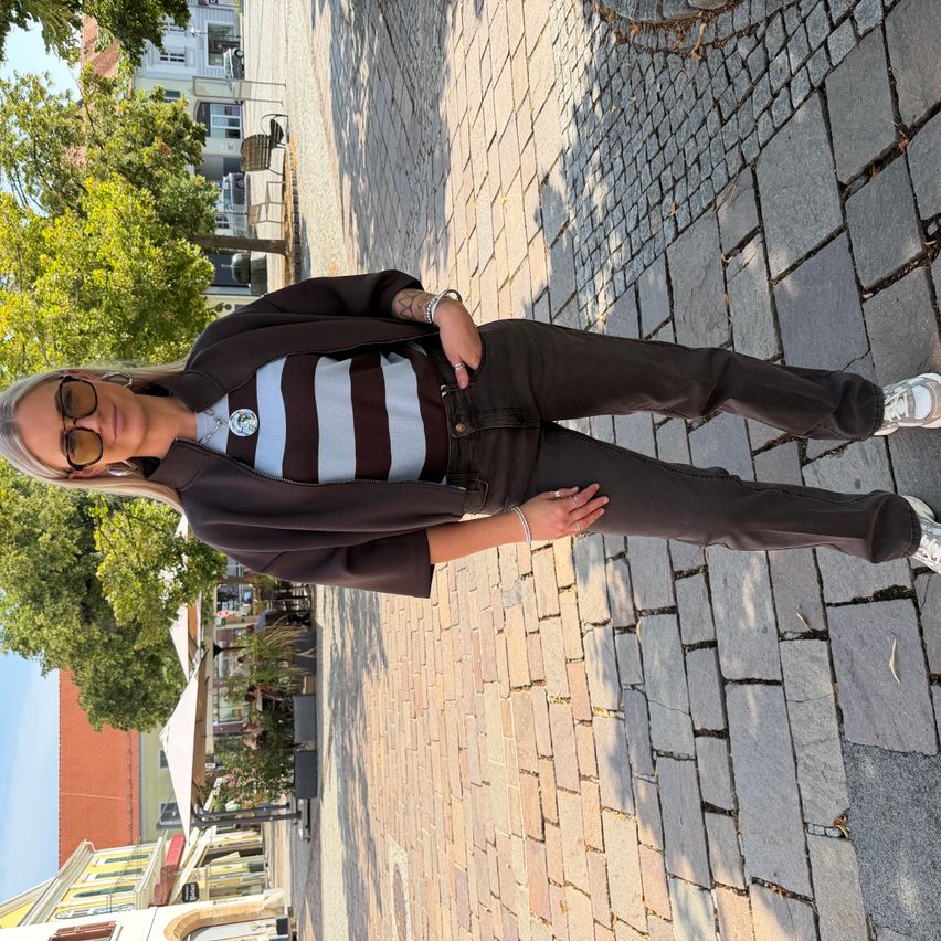 Eine Frau in einem gestreiften Shirt und braunen Hosen posiert auf einer Kopfsteinpflasterstraße. Sie hat ein Tattoo am Arm und trägt eine Sonnenbrille.