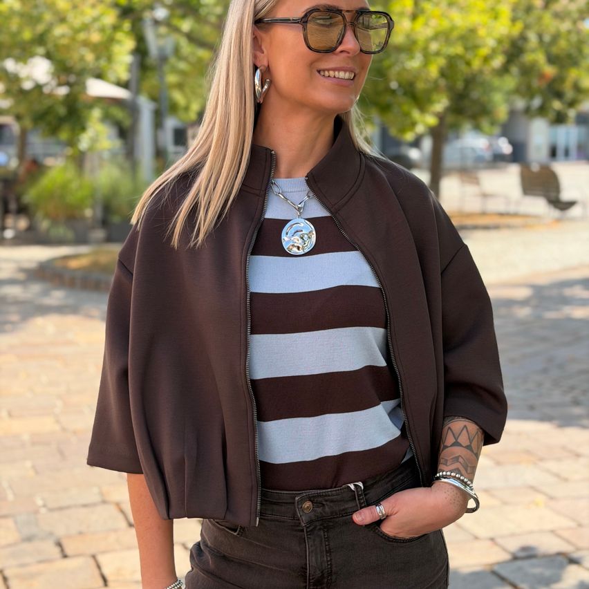 Eine Frau mit blonden Haaren, die eine braune Jacke und ein gestreiftes Shirt trägt, steht auf einem gepflasterten Weg mit Bäumen im Hintergrund.
