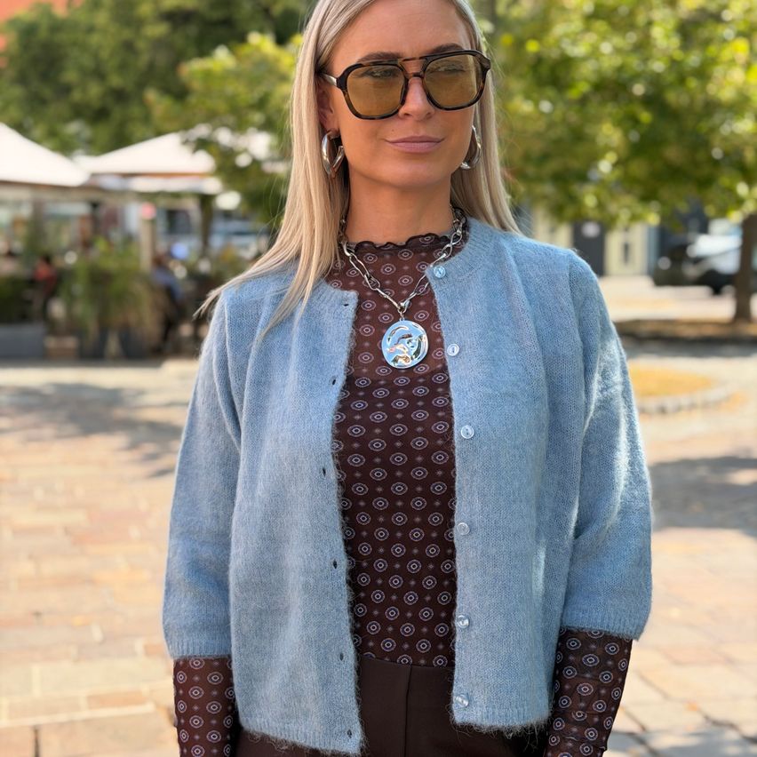 Eine Frau mit blonden Haaren trägt einen hellblauen Cardigan, ein gemustertes Hemd und eine Sonnenbrille auf einem gepflasterten Bürgersteig.