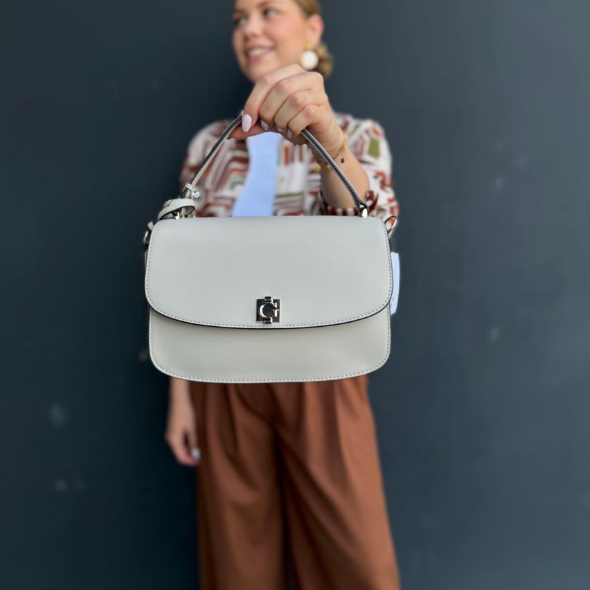 Eine Frau in braunen Hosen hält eine hellgraue Handtasche mit silbernem Logo vor dunklem Hintergrund.