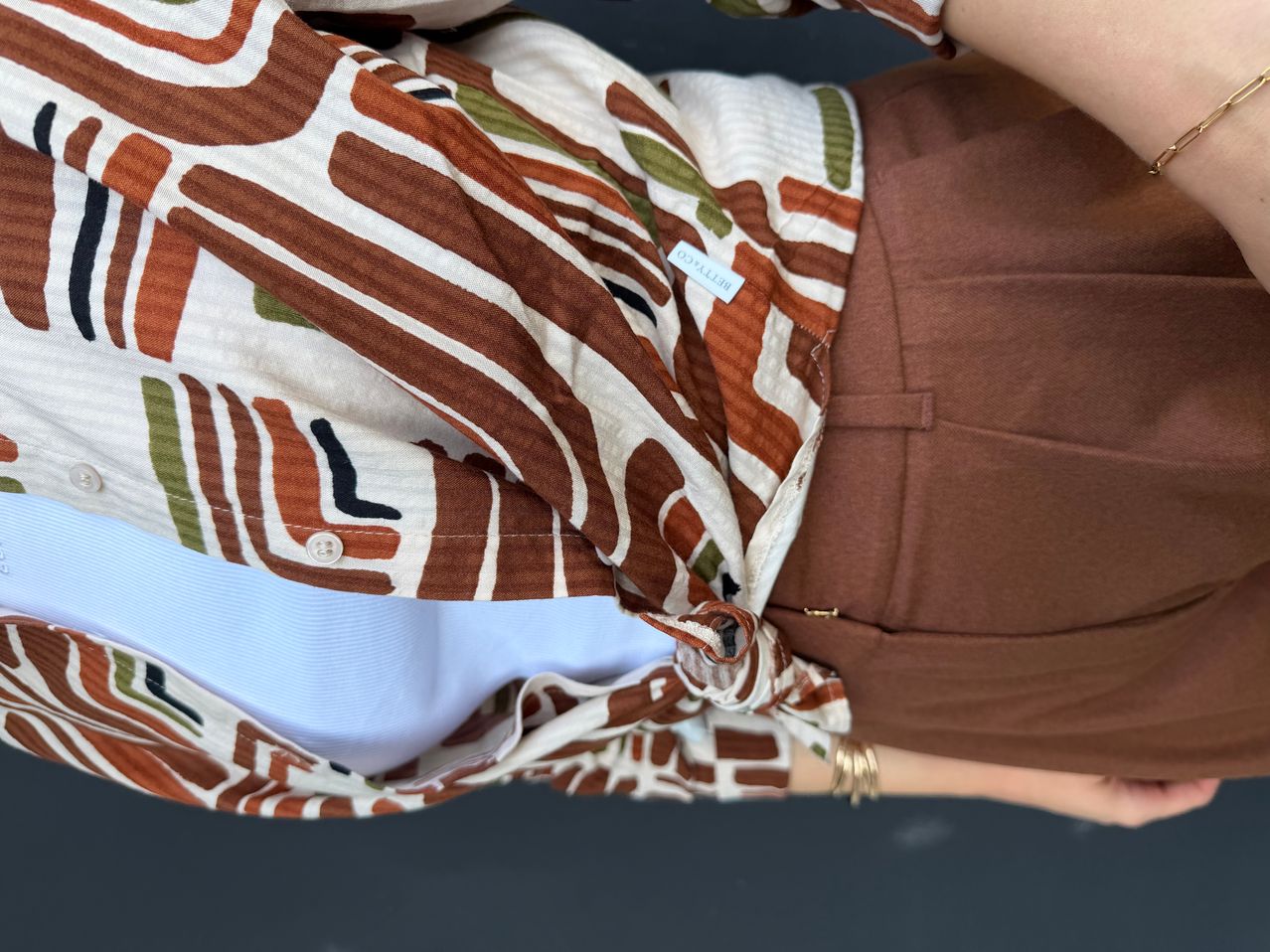 Nahaufnahme des Oberkörpers einer Person, die ein gemustertes Hemd mit braunen, weißen und grünen Streifen trägt. Das Hemd ist um die Taille gebunden.