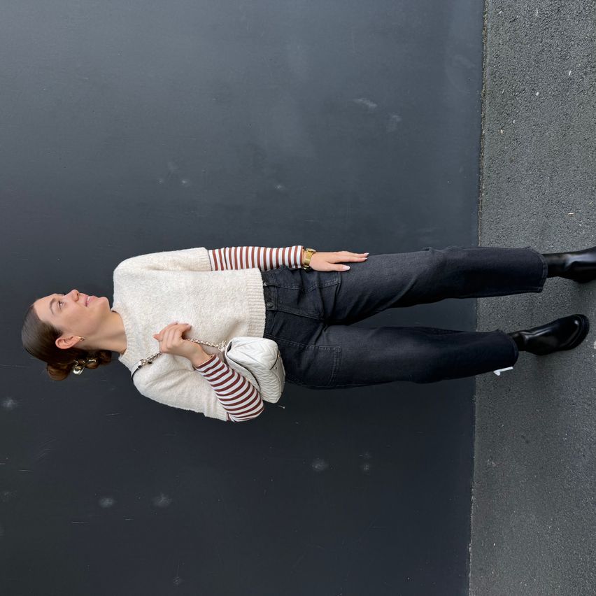 Eine Frau in einem weißen Pullover mit roten und weißen Streifen, schwarzen Jeans und schwarzen Stiefeln liegt auf einem dunklen Hintergrund.