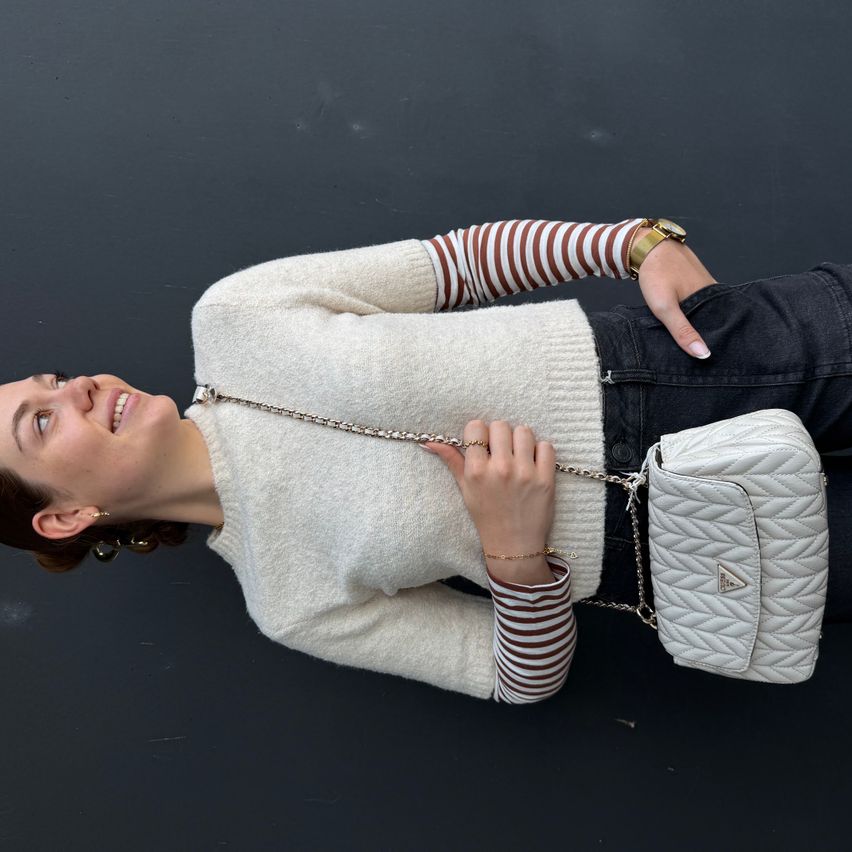Eine Frau liegt auf einer schwarzen Oberfläche, trägt einen weißen Pullover, ein gestreiftes Hemd und hält eine weiße gesteppte Tasche.