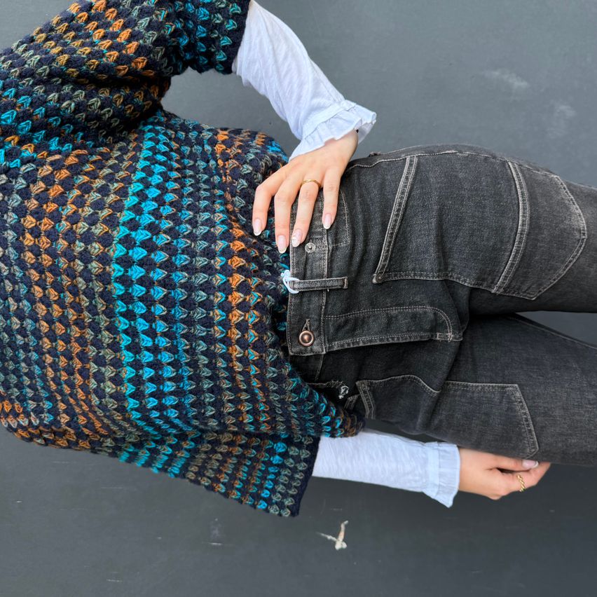 Eine Person, die einen bunten gestrickten Pullover und Jeans trägt, liegt auf einer grauen Oberfläche. Der Pullover hat ein geometrisches Muster mit blauen, orangefarbenen und grünen Farben.