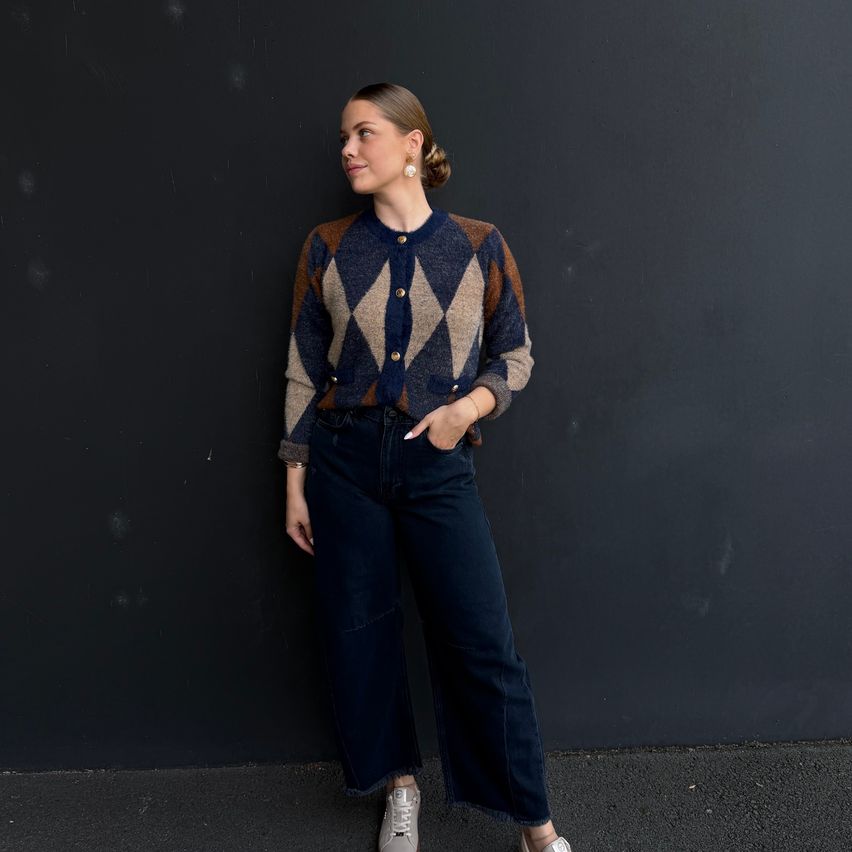 A woman stands against a black wall wearing a patterned cardigan, blue jeans, and white sneakers. She has a serious expression and her hands are in her pockets.