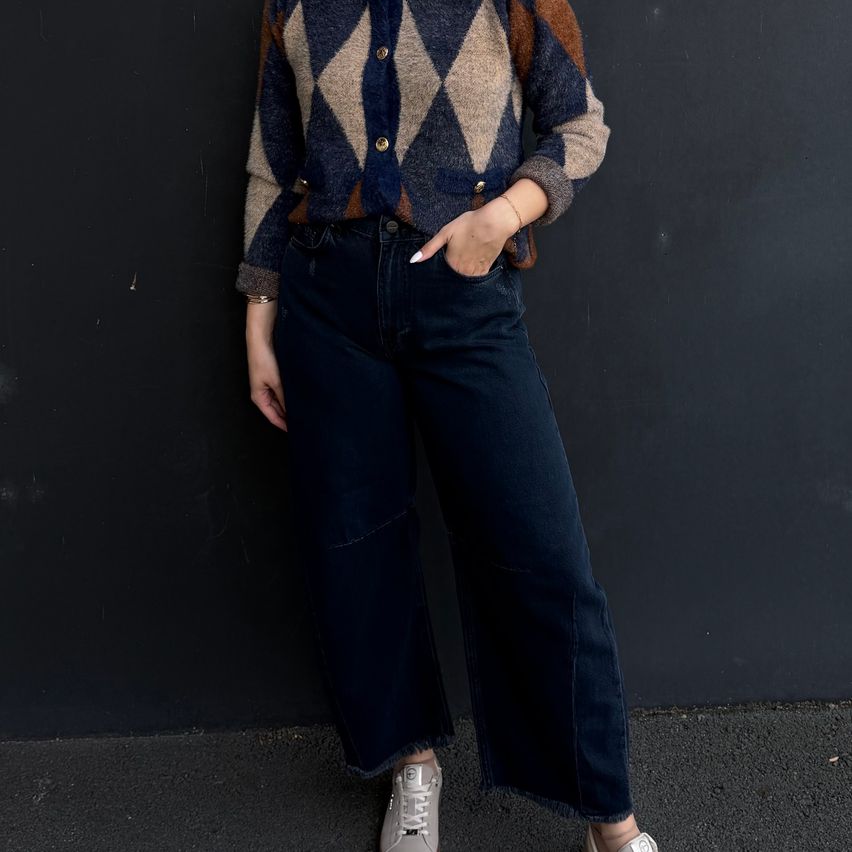 A woman wearing a blue and brown argyle sweater with a bracelet stands in front of a black wall.