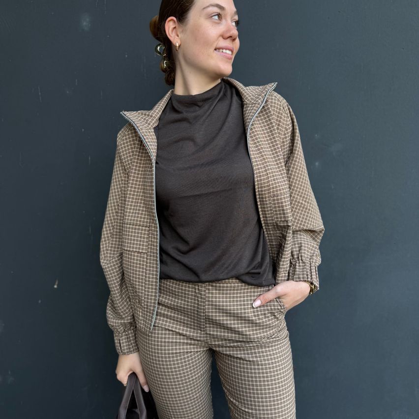 Eine Frau in einem schwarzen Rollkragenpullover und einer braun karierten Jacke mit passender Hose steht gegen eine graue Wand und lächelt.