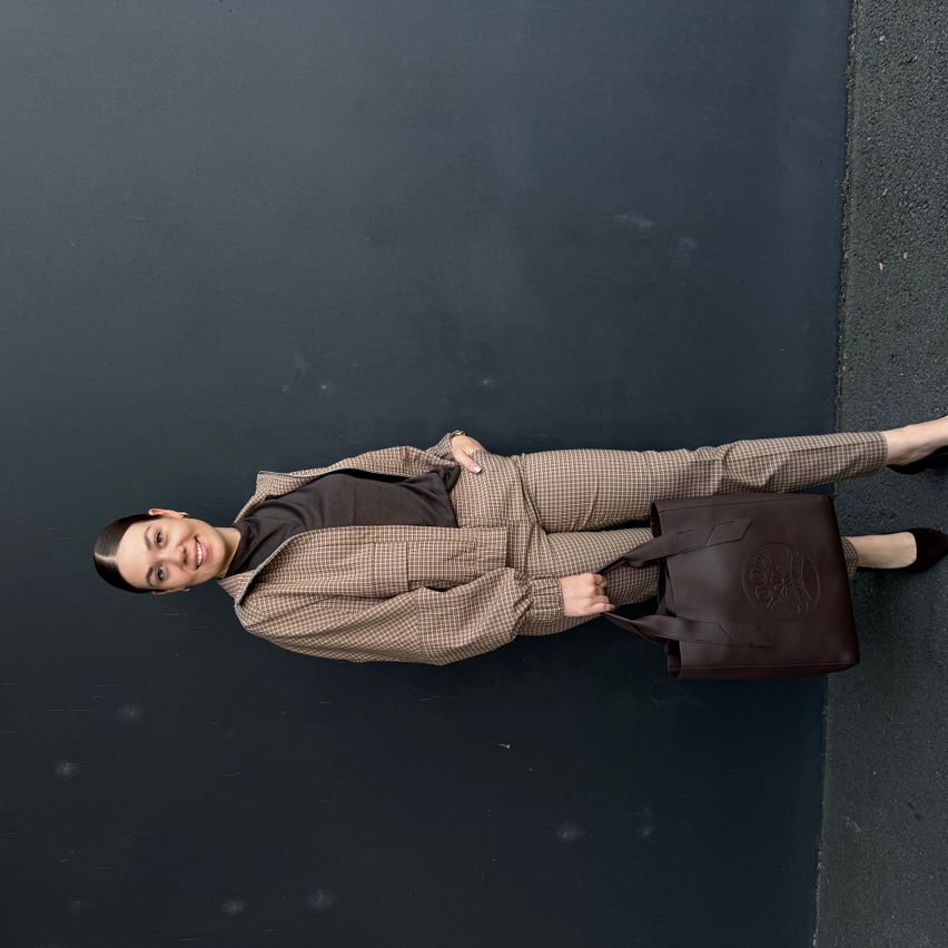 Eine Frau in einem braunen Karoanzug hält eine braune Ledertasche, während sie auf einem schwarzen Hintergrund liegt.