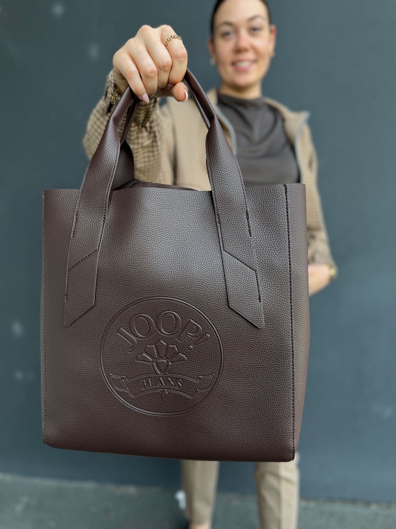 Eine Frau hält eine braune Leder-Tote-Bag mit dem geprägten Logo 'JOOP! JEANS' auf der Vorderseite. Die Tasche hat ein einfaches Design mit zwei Henkeln.