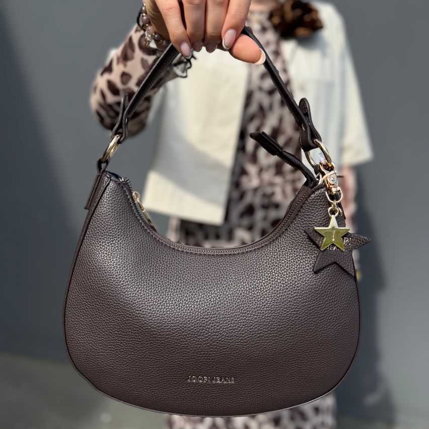 Eine Frau hält eine braune Lederhandtasche mit einem Sternanhänger. Die Tasche hat einen geschwungenen Griff und wird von einer Person in einem Leopardenmusterkleid gehalten.