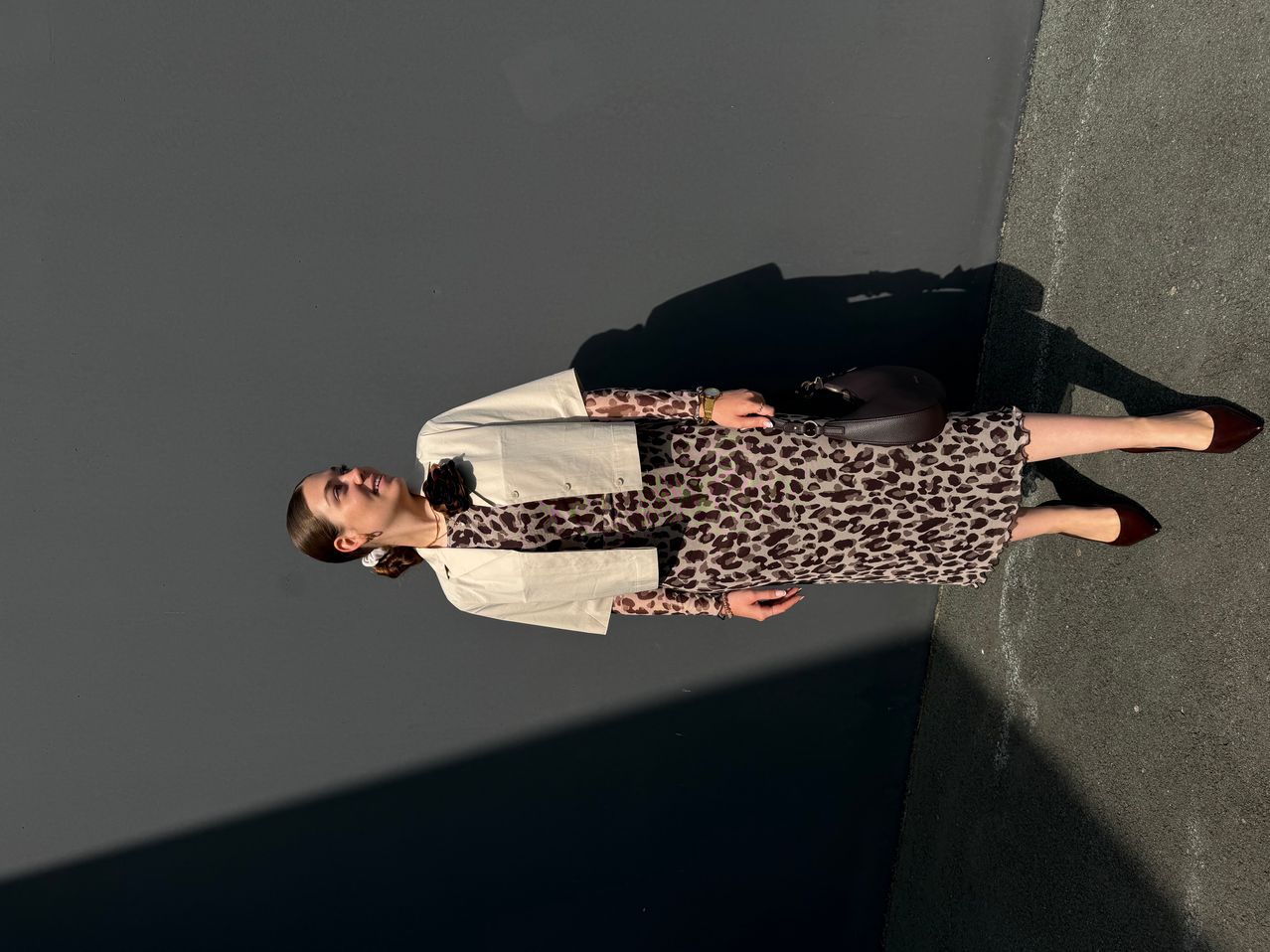 Eine Frau in einem Leopardenmusterkleid und einer beigefarbenen Jacke steht auf einer grauen Wand, lächelt und hält eine braune Handtasche.