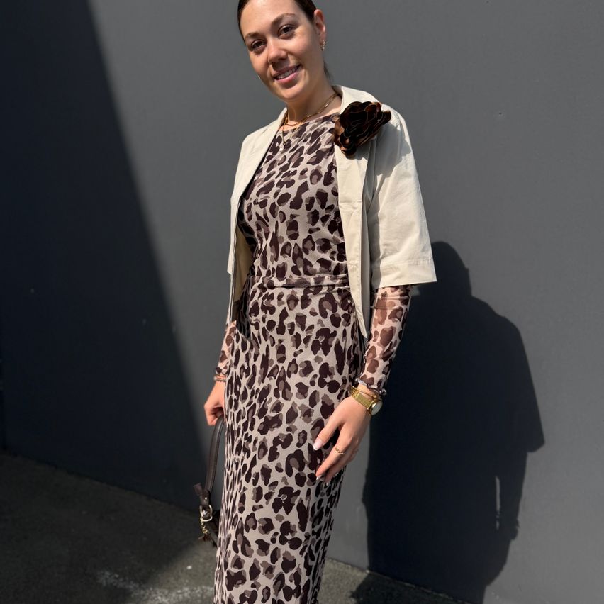 Eine Frau in einem Leopardenmusterkleid und einer weißen Jacke steht gegen eine graue Wand, lächelt und hält eine Handtasche.