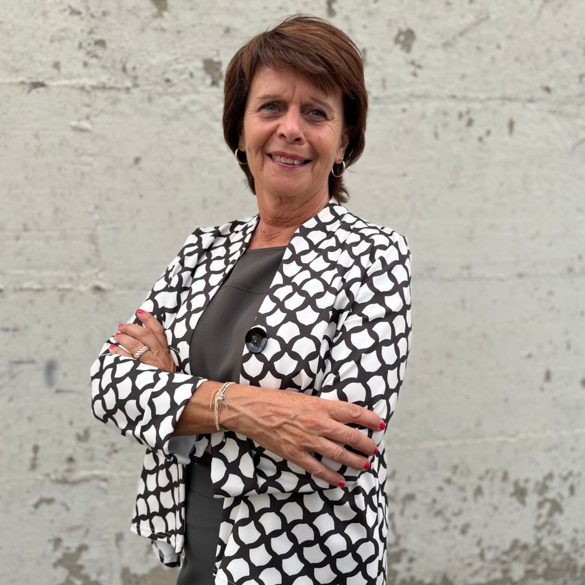 A smiling woman with short hair, wearing a black and white patterned blazer, stands with her arms crossed against a textured concrete wall.
