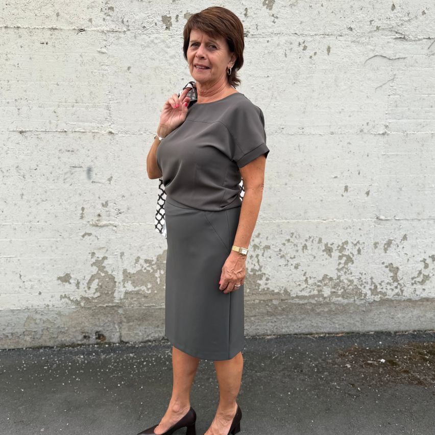 An older woman stands against a white wall, wearing a gray skirt and top, holding a black and white bag. She has short hair and is smiling.
