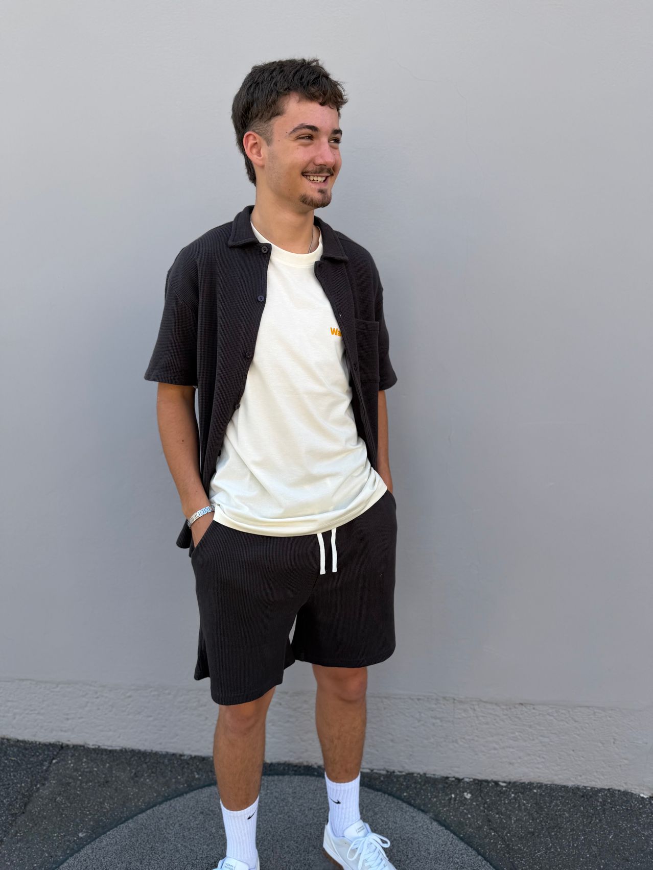 Ein Mann in einem weißen T-Shirt und schwarzen Shorts, der mit einem Lächeln auf dem Gesicht gegen eine schlichte Wand lehnt.