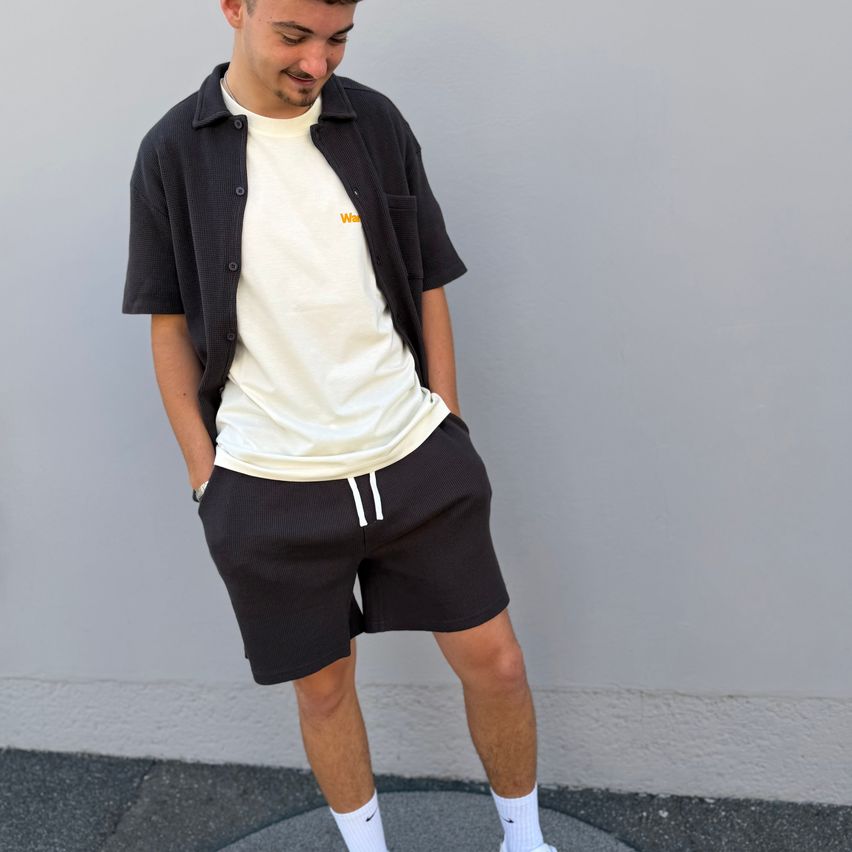 Ein Mann in einem weißen T-Shirt und schwarzen Shorts steht mit den Händen in den Taschen an einer grauen Wand.