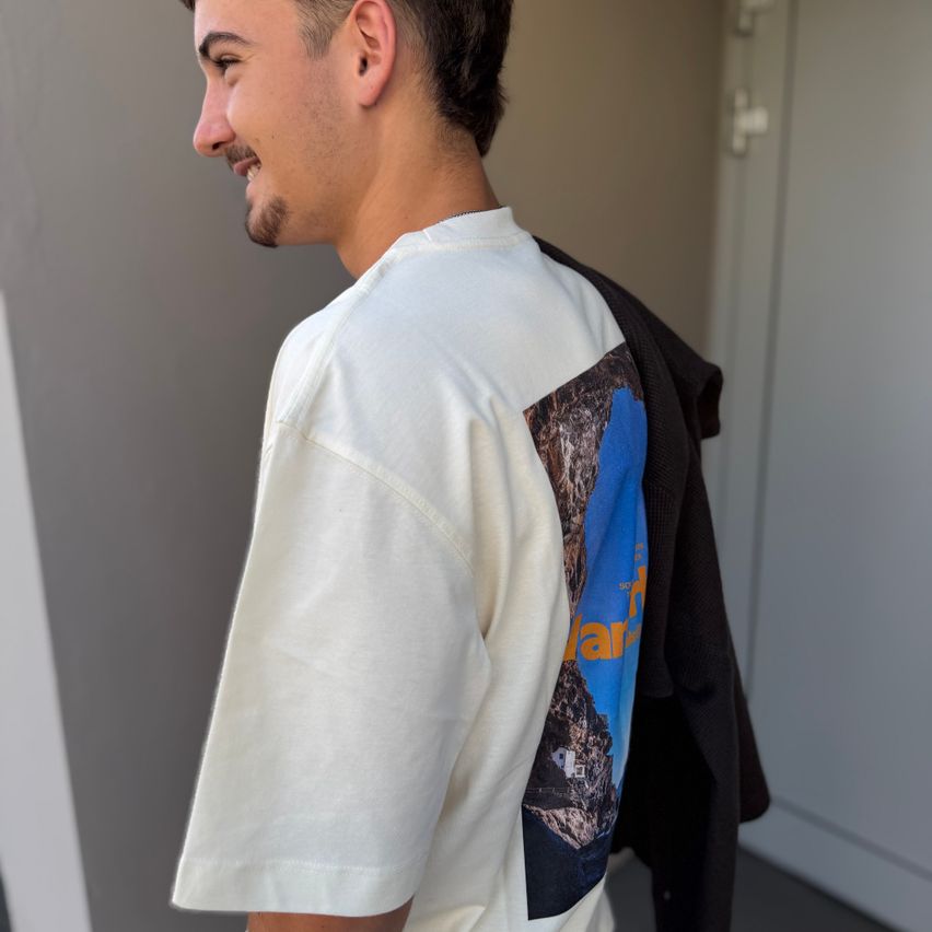 Ein Mann mit Bart trägt ein weißes T-Shirt mit einem blauen und orangefarbenen Grafikdesign auf dem Rücken. Er steht vor einer grauen Wand.