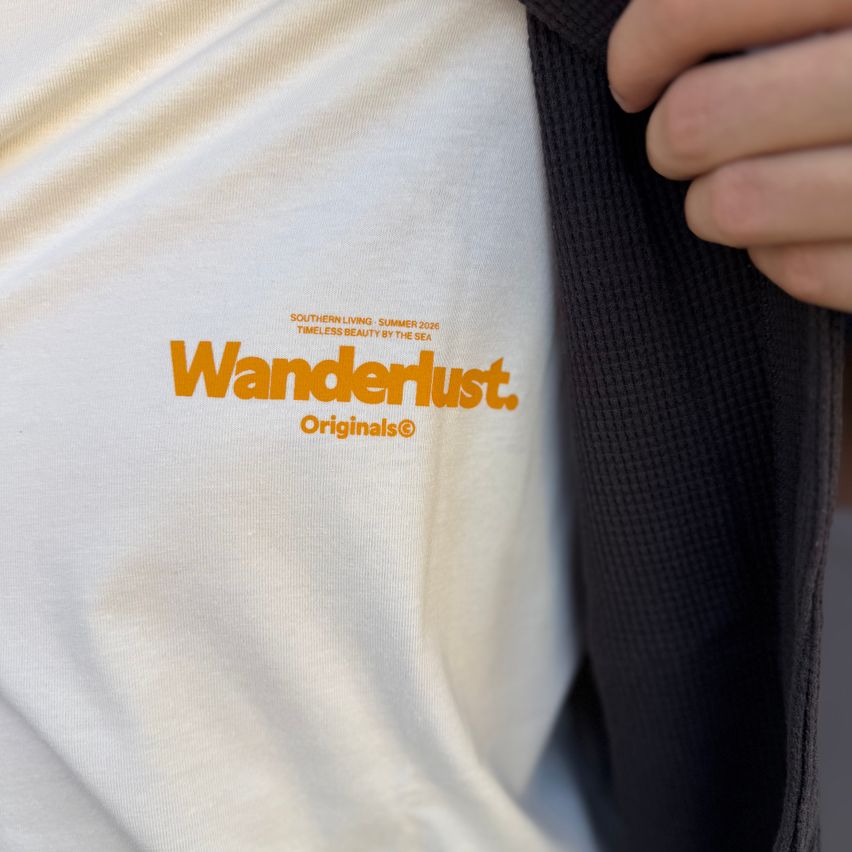 Nahaufnahme eines weißen T-Shirts mit orangefarbenem Text 'Wanderlust. Originals' auf der linken Brust. Das Shirt sieht getragen aus und hat einen leichten Fleck.