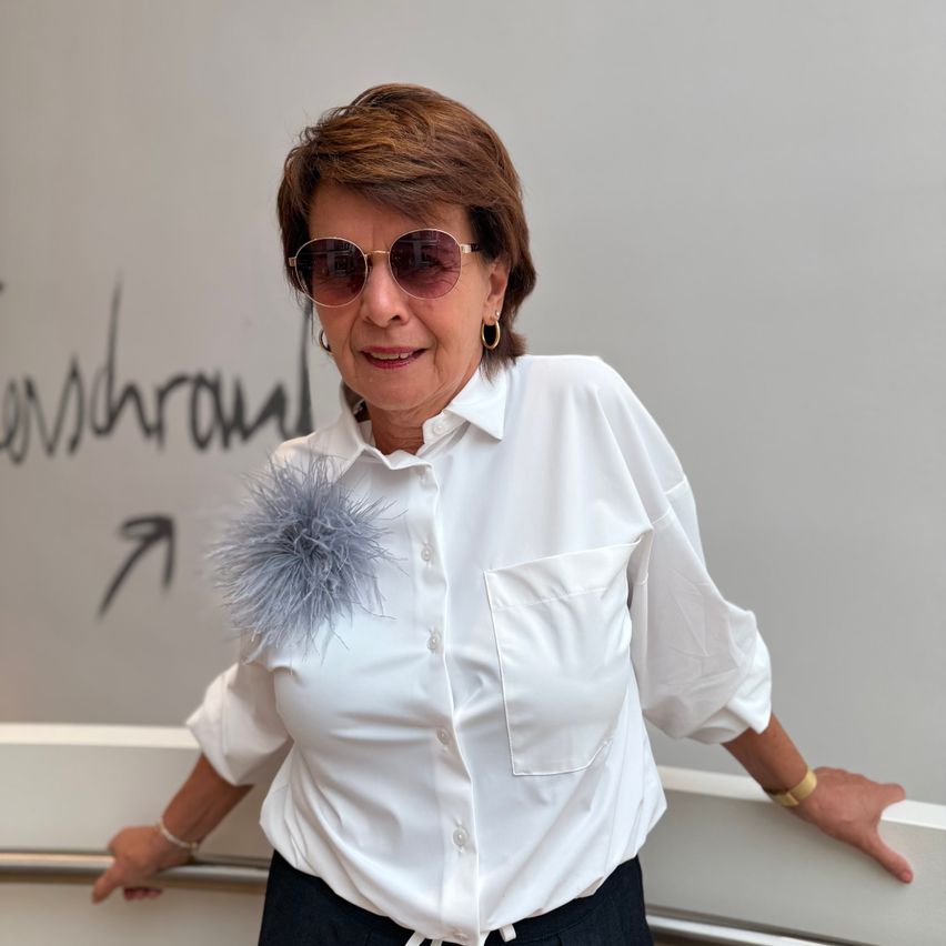 A woman with short hair wearing a white shirt with a feather accessory on the shoulder, sunglasses, and a bracelet poses in front of a wall with a logo.