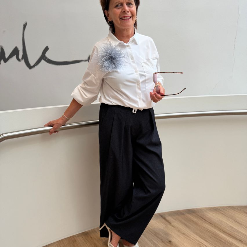 A woman in a white shirt and black pants stands on a balcony, smiling and holding a pair of glasses.
