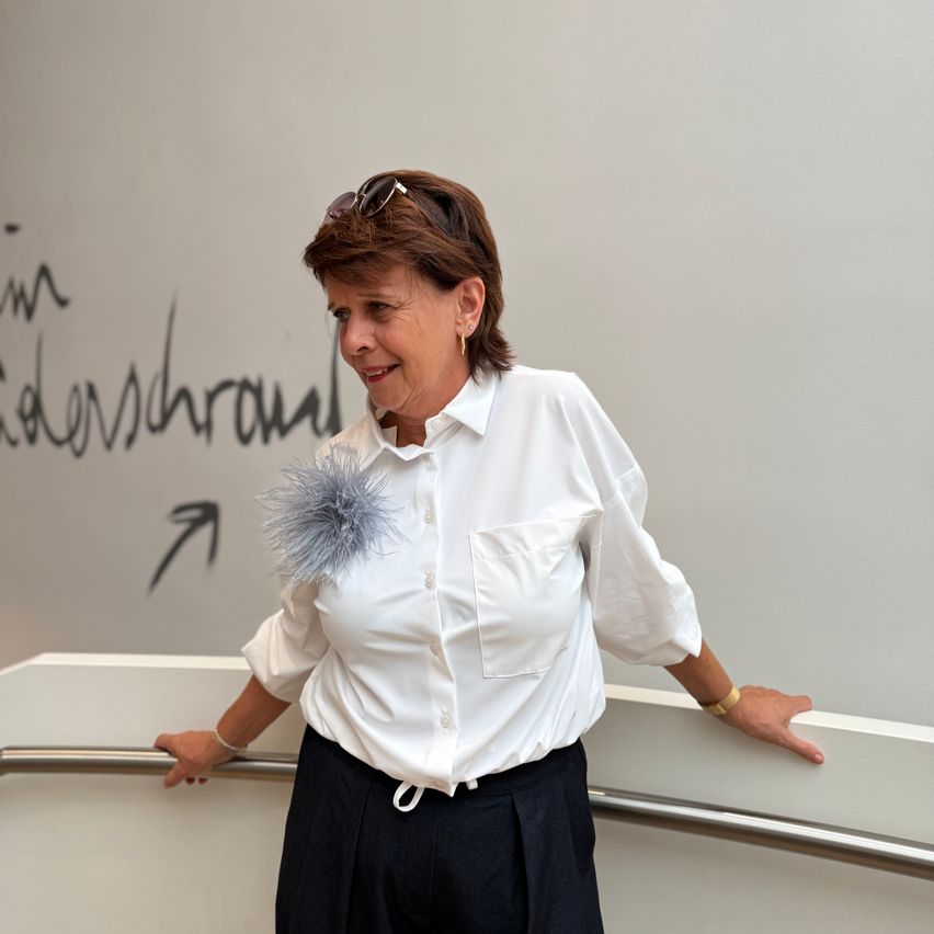 A woman with a gray feather accessory on her shirt stands by a white wall with graffiti. She is smiling and leaning on a handrail.