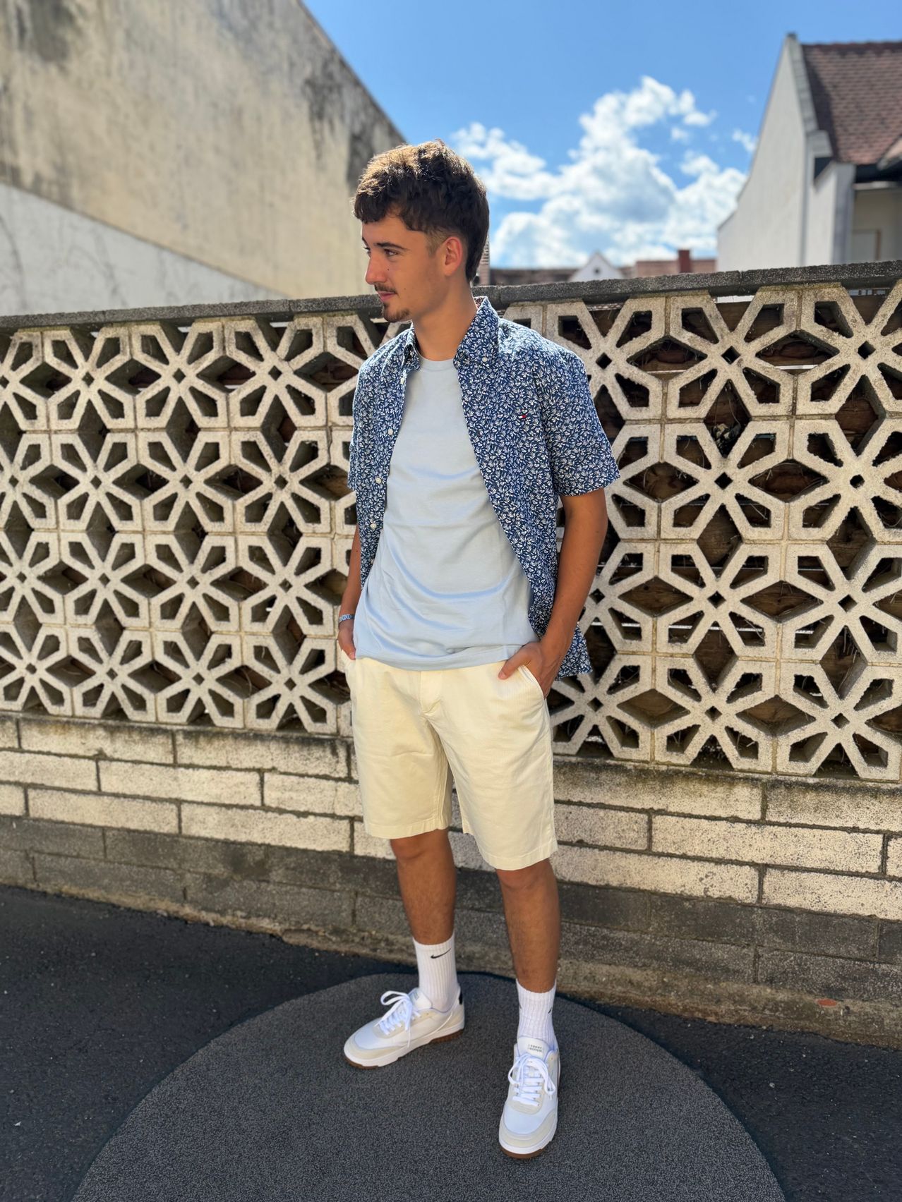 A man stands in front of a decorative wall, wearing a light blue shirt and cream shorts. He has his hands in his pockets and is looking to the side.