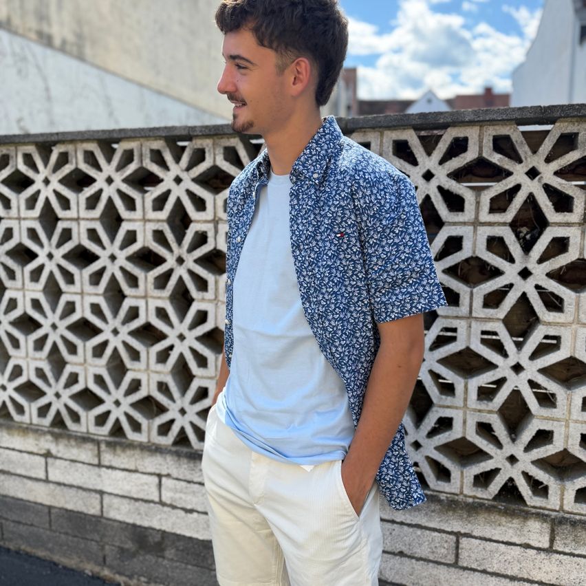 A man in a blue floral shirt and white pants stands against a patterned wall, smiling.
