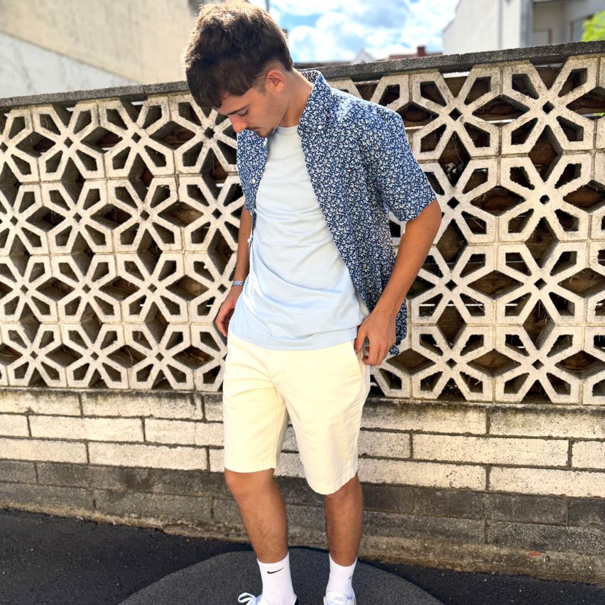 A young man wearing a light blue shirt and cream-colored shorts stands on a sidewalk, looking down. He is next to a decorative concrete wall.