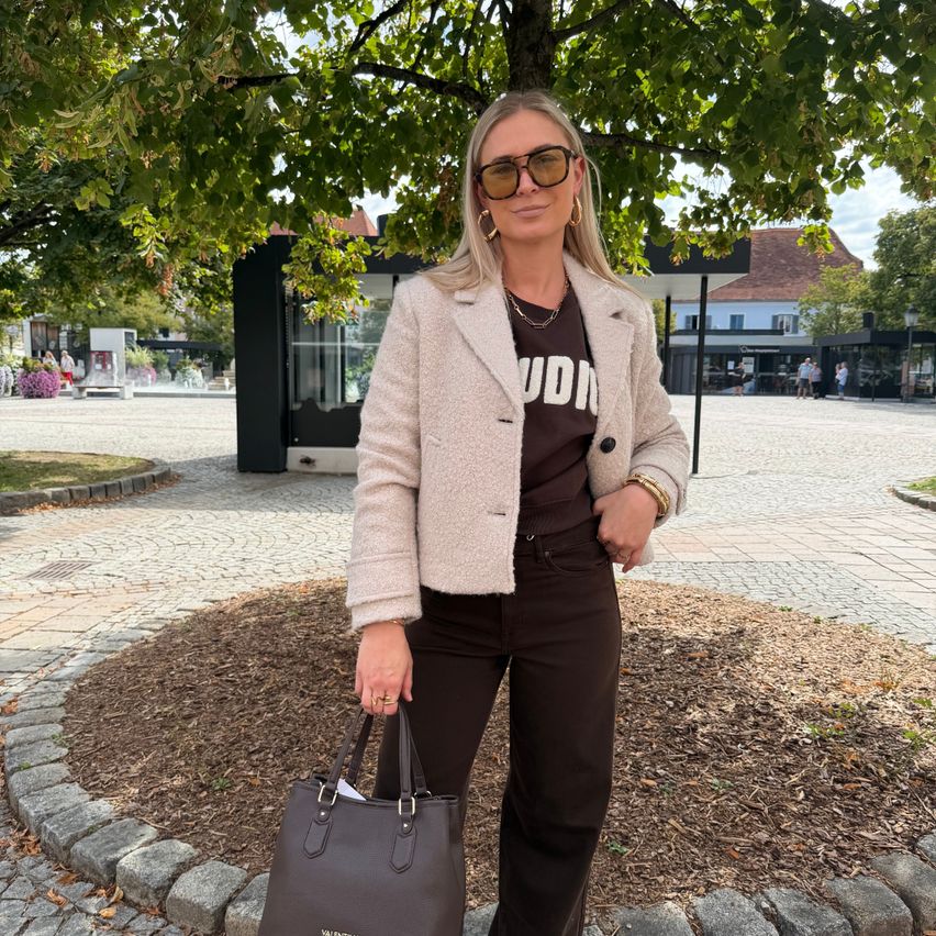 A woman with blonde hair wearing a beige coat, black t-shirt, and brown pants stands in a park with a handbag.