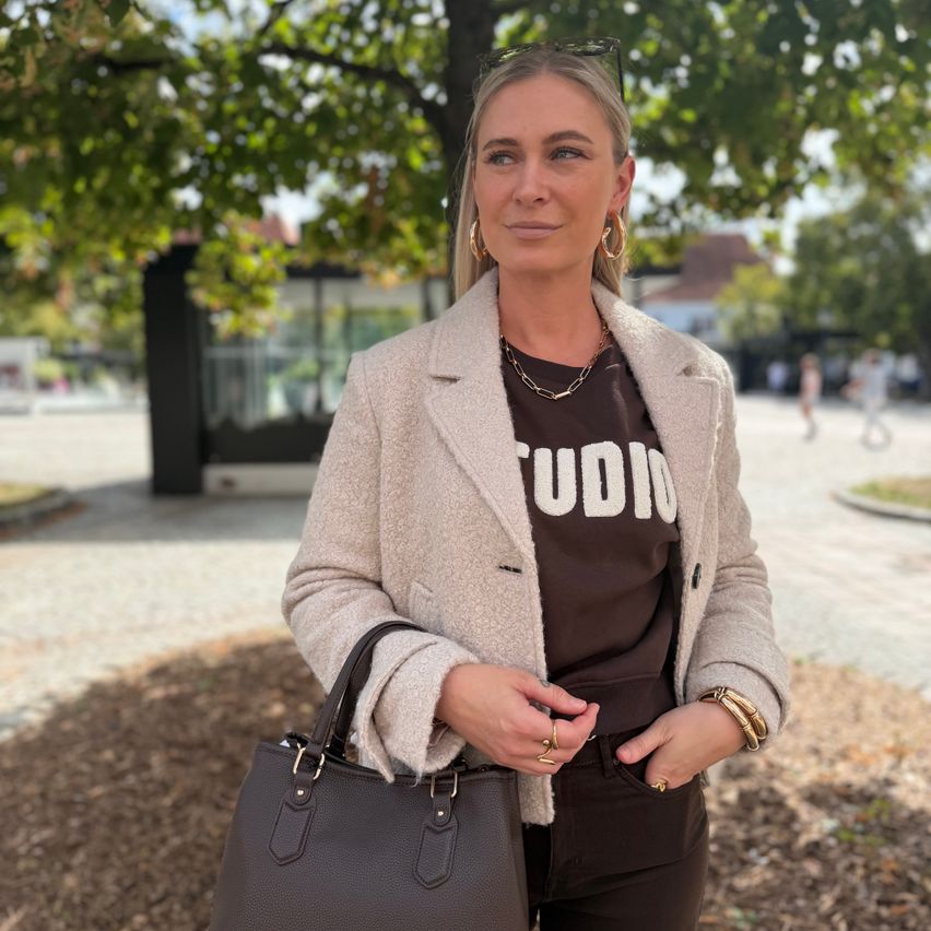 A woman with blonde hair wearing a brown shirt with the word Studio on it, a beige coat, and black pants is standing outside.