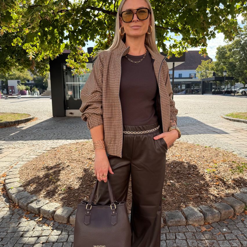 A woman stands under a tree, wearing a brown plaid jacket, brown top, and brown leather pants. She holds a brown handbag and has a necklace and bracelets.