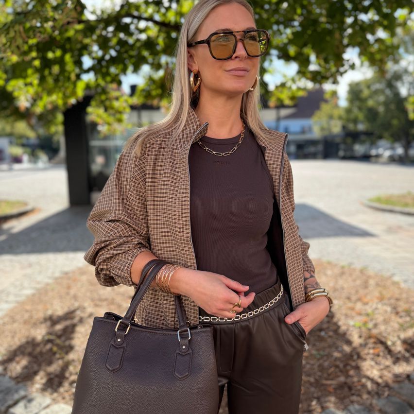 A woman with blonde hair is standing outdoors, wearing sunglasses, a necklace, and a checkered jacket. She holds a brown handbag and has a ring on her finger.