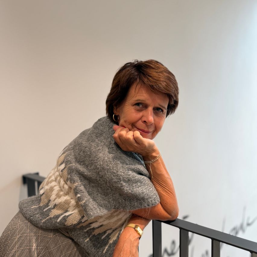 A woman with short hair, wearing a gray shawl, is standing on a staircase and smiling at the camera.