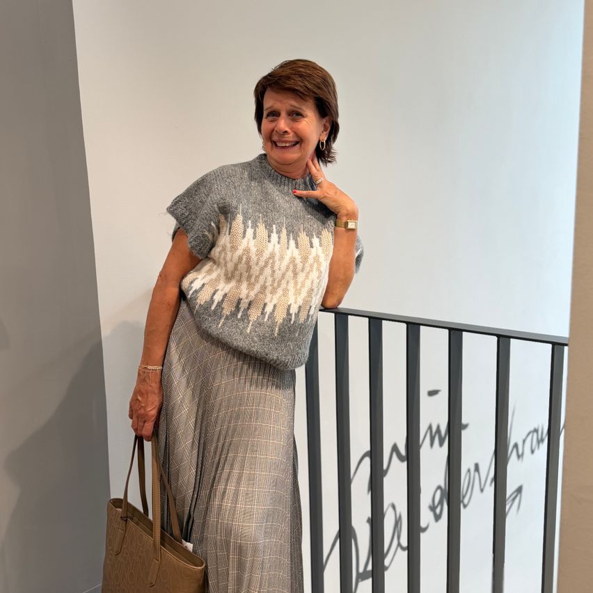 A woman stands on a staircase, smiling, wearing a patterned sweater and plaid skirt, holding a brown handbag. Behind her is a white wall with black railings.