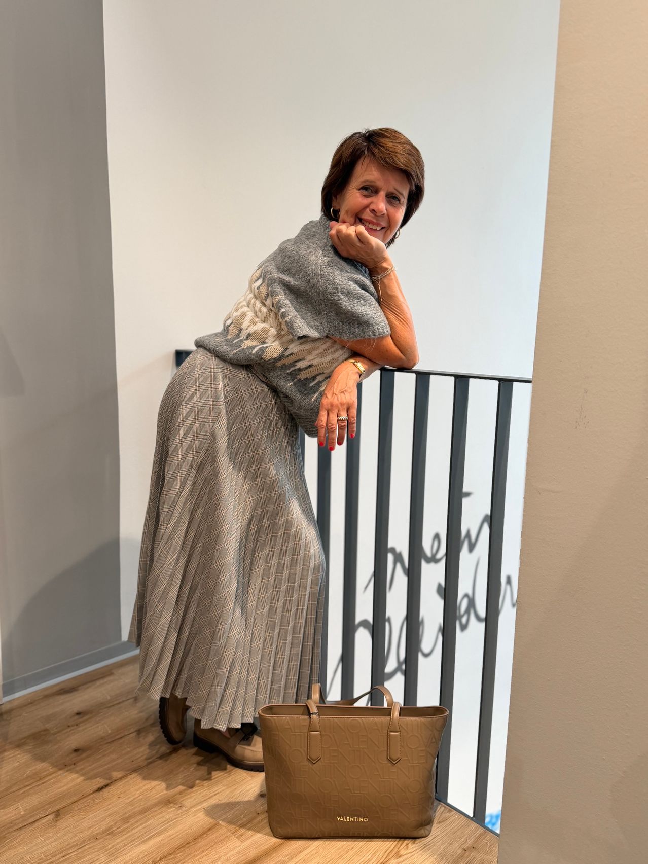 A woman in a gray sweater and a pleated skirt stands on a balcony, smiling and leaning on the railing. A brown handbag is placed on the floor.