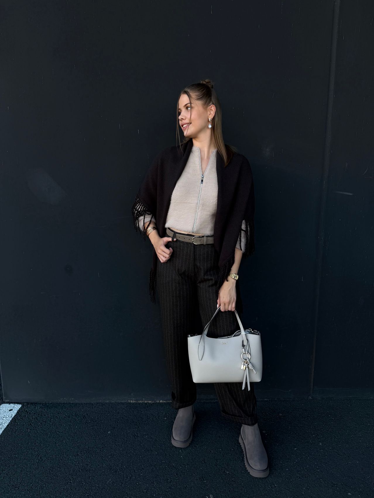 A woman stands against a black wall, smiling, wearing a gray top, black pants, and a black fringed shawl, holding a white handbag.
